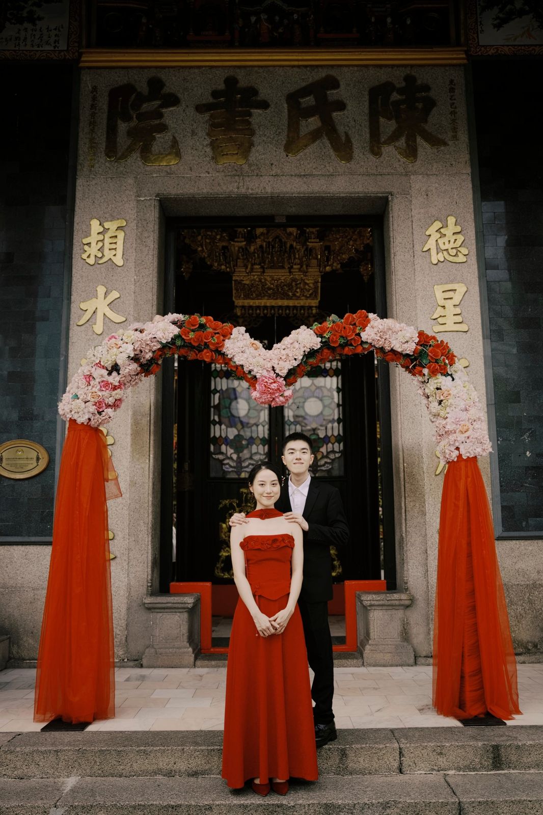 The couple standing under a flower arch in front of the hall
